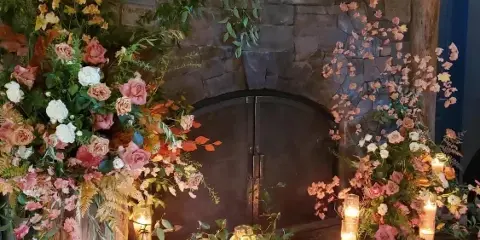 A stone fireplace decorated with lush floral arrangements, greenery, and lit candles.