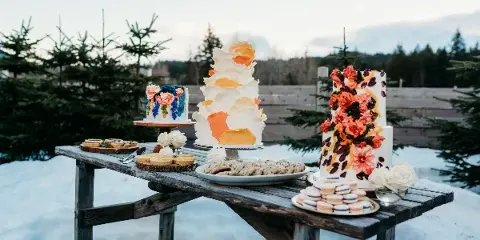A rustic wooden table outdoors in a snowy setting displays several elaborately decorated cakes, including a tall white cake with orange accents and a floral cake, alongside plates of macarons and cookies.