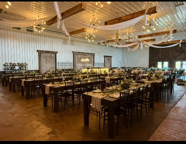 Interior of a rustic barn wedding venue featuring exposed wooden beams, chandeliers, and long dining tables set for a reception.