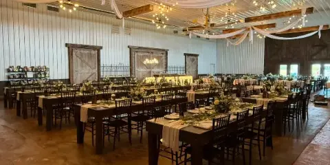 Interior of a rustic barn wedding venue featuring exposed wooden beams, chandeliers, and long dining tables set for a reception.