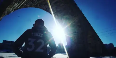 Cinematic low-angle shot of a basketball player wearing a jersey with the name 'TOWNS' and number 32, standing under a bridge with bright sun flare.