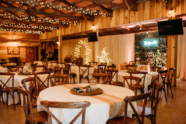 Interior view of a rustic barn wedding venue with wooden beams, string lights, and round tables set with white linens and wooden chairs.