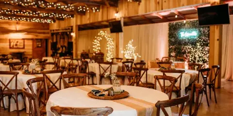 Interior view of a rustic barn wedding venue with wooden beams, string lights, and round tables set with white linens and wooden chairs.
