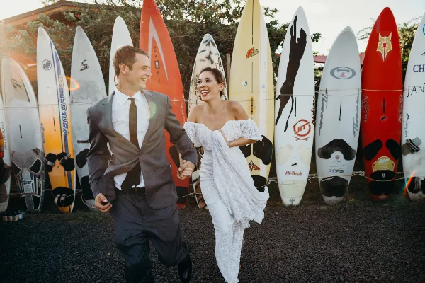 A bride and groom laughing and holding hands while walking in front of a row of surfboards.