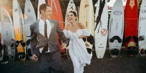A bride and groom laughing and holding hands while walking in front of a row of surfboards.