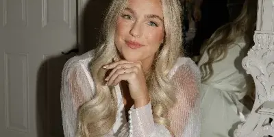 A blonde bride with professional makeup and styled hair posing in a white robe in front of a mirror.