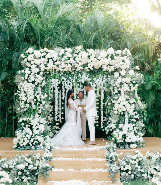 A bride and groom exchanging vows under a large white floral arch in a tropical outdoor setting.