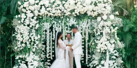 A bride and groom exchanging vows under a large white floral arch in a tropical outdoor setting.