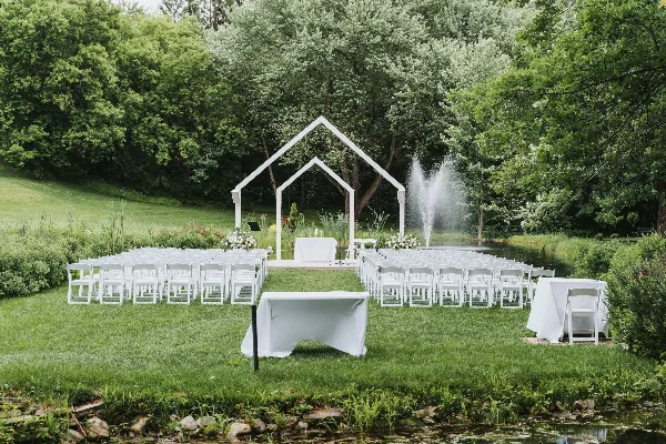 An outdoor wedding ceremony setup with rows of white folding chairs and white-draped tables arranged on a lawn near a pond.