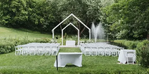 An outdoor wedding ceremony setup with rows of white folding chairs and white-draped tables arranged on a lawn near a pond.
