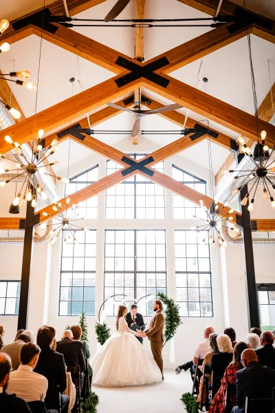 Wedding ceremony in a modern rustic venue with high ceilings, exposed wooden beams, and large windows.