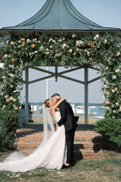 A bride and groom sharing a kiss under a floral-decorated gazebo arch at a waterfront wedding venue.