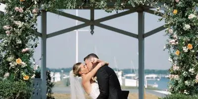A bride and groom sharing a kiss under a floral-decorated gazebo arch at a waterfront wedding venue.