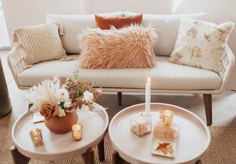 A styled vintage living room scene featuring a rattan sofa with textured pillows, a floral arrangement in a terracotta pot, and candles on a round coffee table.