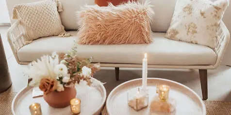 A styled vintage living room scene featuring a rattan sofa with textured pillows, a floral arrangement in a terracotta pot, and candles on a round coffee table.