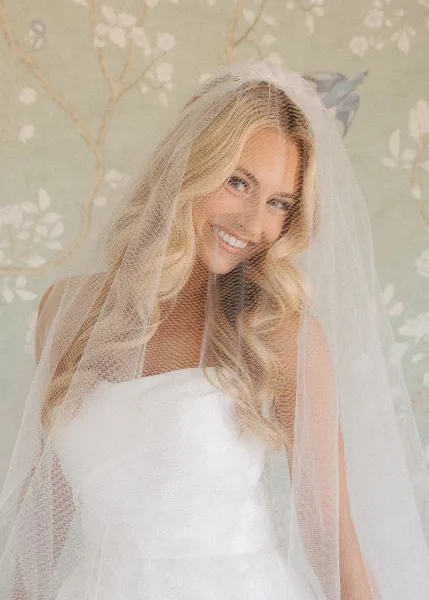 A smiling blonde bride wearing a white wedding dress and veil, showcasing styled hair and makeup.