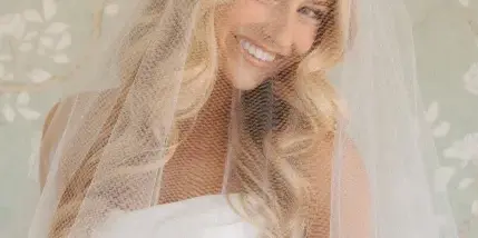 A smiling blonde bride wearing a white wedding dress and veil, showcasing styled hair and makeup.