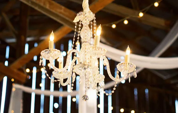 A lit crystal chandelier hanging in a rustic event venue with wooden beams and string lights.