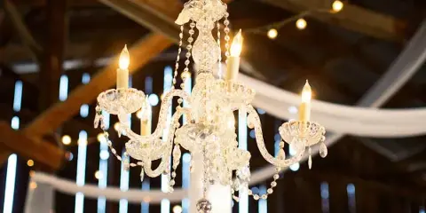 A lit crystal chandelier hanging in a rustic event venue with wooden beams and string lights.