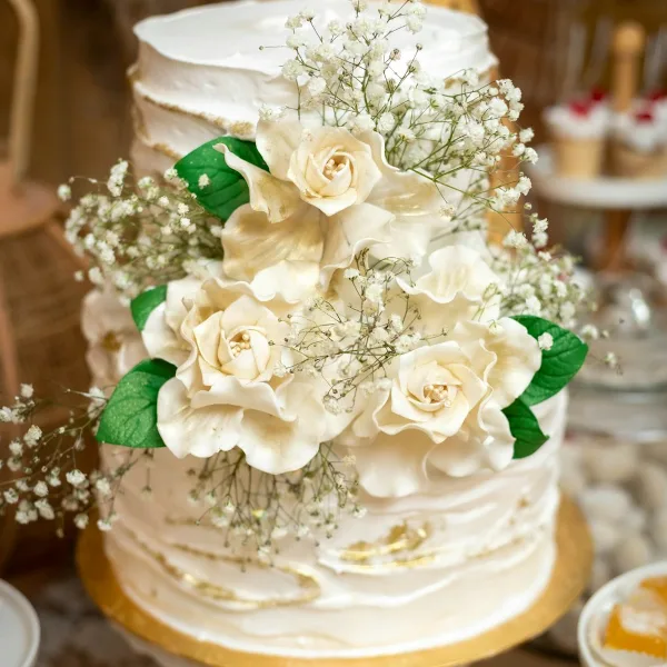 A close-up of a white wedding cake decorated with cream-colored sugar roses, green leaves, and baby's breath, featuring rustic frosting and gold accents.