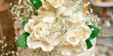 A close-up of a white wedding cake decorated with cream-colored sugar roses, green leaves, and baby's breath, featuring rustic frosting and gold accents.