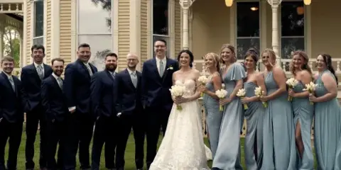 A wedding party consisting of a bride, groom, and attendants posing for a group photo in front of a venue.