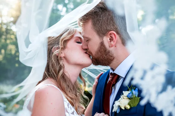 A close-up professional photograph of a bride and groom kissing under a veil in an outdoor setting.