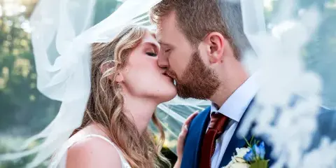 A close-up professional photograph of a bride and groom kissing under a veil in an outdoor setting.
