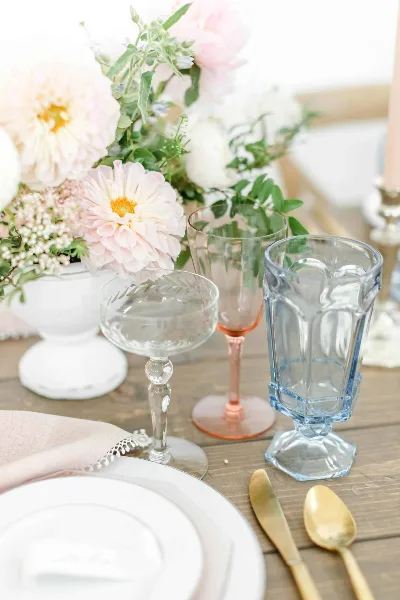 A close-up of a table setting with mixed vintage glassware, gold cutlery, and a floral arrangement.