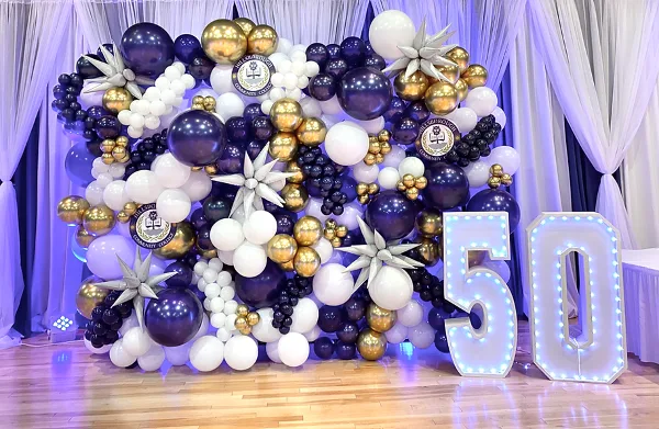 A large event balloon arch backdrop in purple, gold, and white, featuring star-shaped balloon accents and illuminated marquee numbers '50' on a wooden floor.