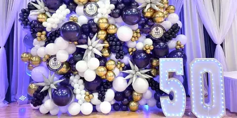 A large event balloon arch backdrop in purple, gold, and white, featuring star-shaped balloon accents and illuminated marquee numbers '50' on a wooden floor.