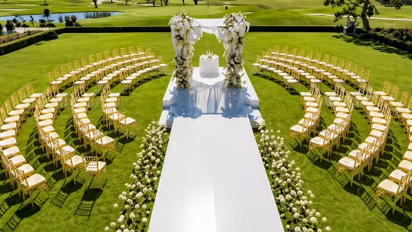 Outdoor wedding ceremony setup featuring rows of gold chiavari chairs, a white floral arch, and a white aisle runner on a green lawn.