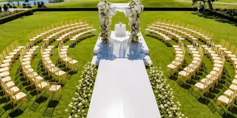 Outdoor wedding ceremony setup featuring rows of gold chiavari chairs, a white floral arch, and a white aisle runner on a green lawn.