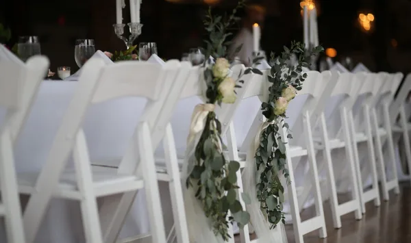 A row of white folding chairs with floral decorations and a table with white linens set for an event.
