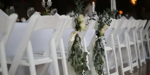 A row of white folding chairs with floral decorations and a table with white linens set for an event.