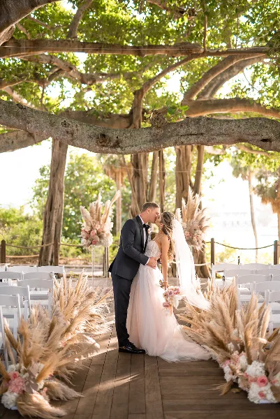 A bride and groom kissing under a large tree at an outdoor wedding ceremony decorated with pampas grass and flowers.