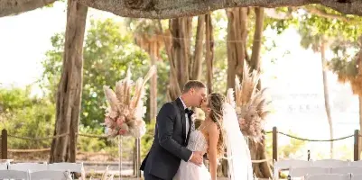 A bride and groom kissing under a large tree at an outdoor wedding ceremony decorated with pampas grass and flowers.