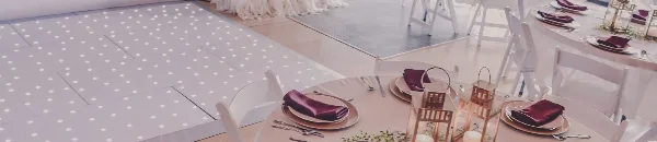 A decorated event reception table with white chairs, place settings, and lanterns next to a dance floor.