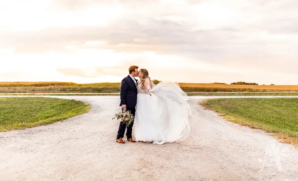 A bride and groom standing on a dirt road in a field at sunset, with the bride's dress blowing in the wind.