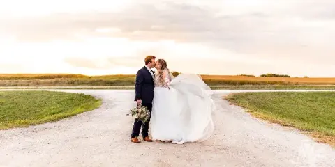 A bride and groom standing on a dirt road in a field at sunset, with the bride's dress blowing in the wind.