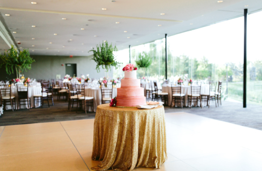 A wedding reception hall featuring a pink tiered cake on a gold tablecloth and set dining tables.