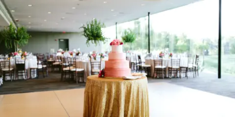 A wedding reception hall featuring a pink tiered cake on a gold tablecloth and set dining tables.