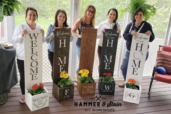 Five women holding up various DIY wooden signs and planters with painted text and flowers, with the Hammer & Stain logo at the bottom.