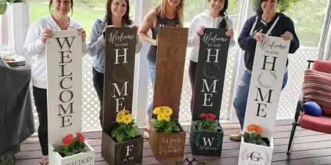 Five women holding up various DIY wooden signs and planters with painted text and flowers, with the Hammer & Stain logo at the bottom.