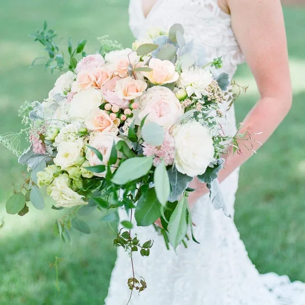 Bride in a white lace wedding dress holding a large bouquet of peach and white roses, hydrangeas, and eucalyptus greenery outdoors on a lawn.