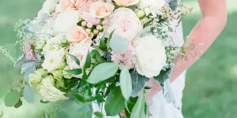 Bride in a white lace wedding dress holding a large bouquet of peach and white roses, hydrangeas, and eucalyptus greenery outdoors on a lawn.