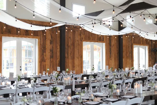 A rustic wooden event hall with white ceiling draping and string lights, featuring long tables set with white chairs, glassware, and floral centerpieces.