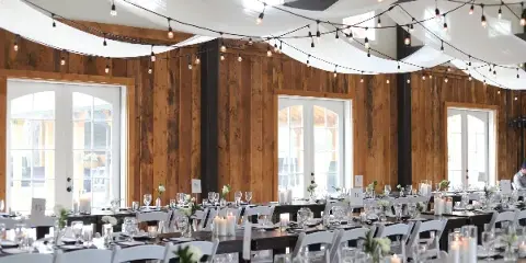 A rustic wooden event hall with white ceiling draping and string lights, featuring long tables set with white chairs, glassware, and floral centerpieces.