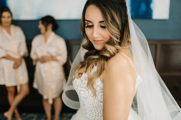A bride with styled hair and makeup looking down, with bridesmaids in robes in the background.