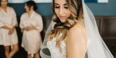 A bride with styled hair and makeup looking down, with bridesmaids in robes in the background.